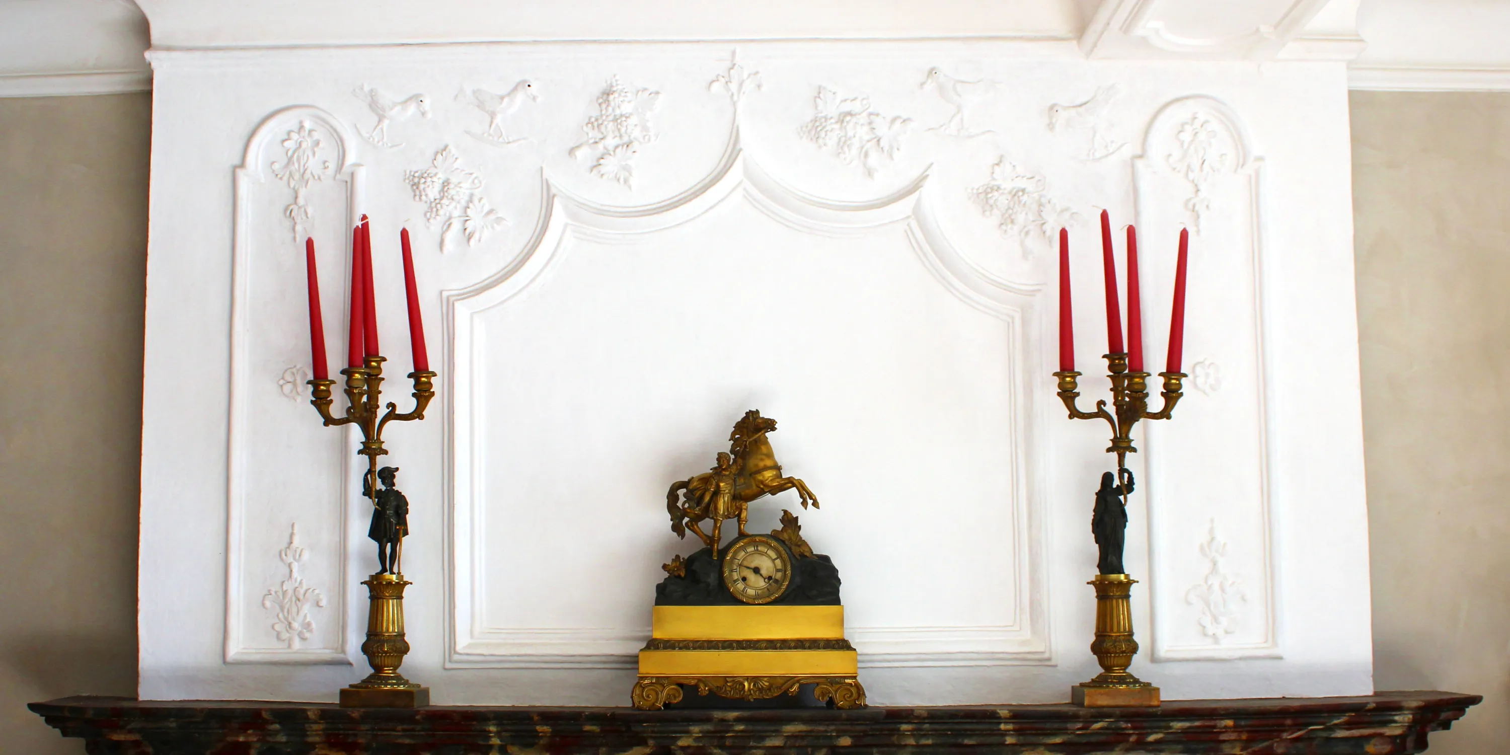 Dans la belle salle de la Blanche Maison, la cheminée de bois peinte en faux marbre est surmontée d’un trumeau en stuc témoignant d’un souci décoratif plutôt rare dans une ferme ardennaise. Techniquement, ces stucs sont un mélange de moulages et de travail à main levée. Leur saveur provient du mélange entre des motifs populaires comme les oiseaux et d’autres plus classiques comme les fleurons. Trumeau en stuc de cheminée comportant quinze motifs agencés de manière symétrique autour de moulures d’inspiration Louis XIV : quatre oiseaux, quatre pampres, cinq fleurons et deux rosaces. Ⓒ 2024 CC BY-NC 4.0 Gilles Pirlot de Corbion